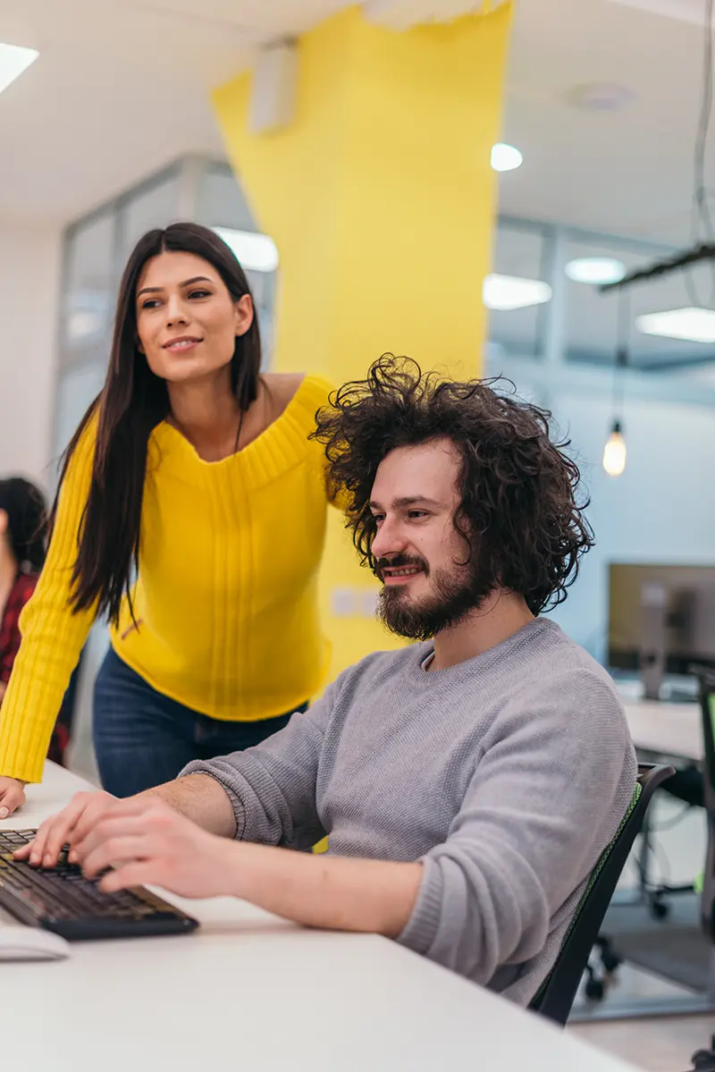 Young man with wild dark hair typing on a keyboard in a modern office with a female co-worker standing alongside wearing a bright yellow jumper.