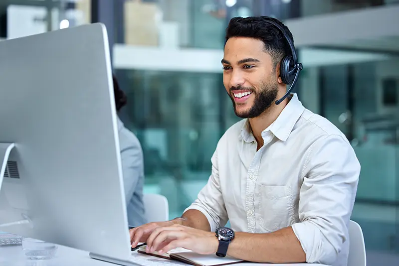 Young male call centre agent looking at a computer monitor, wearing a headset and speaking to a customer.